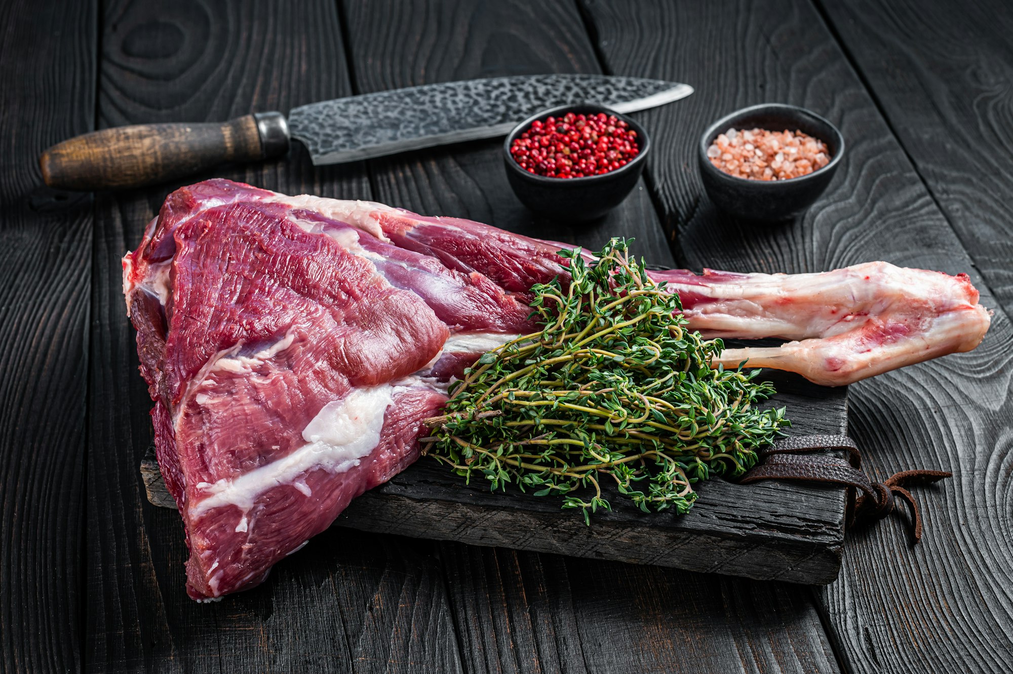 Uncooked Raw goat leg with herbs on butcher cutting board. Black wooden background.