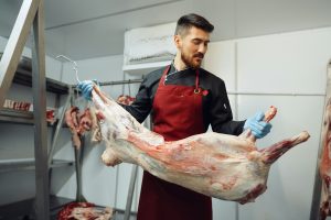 Male butcher standing by meat hanging on hook in meat shop